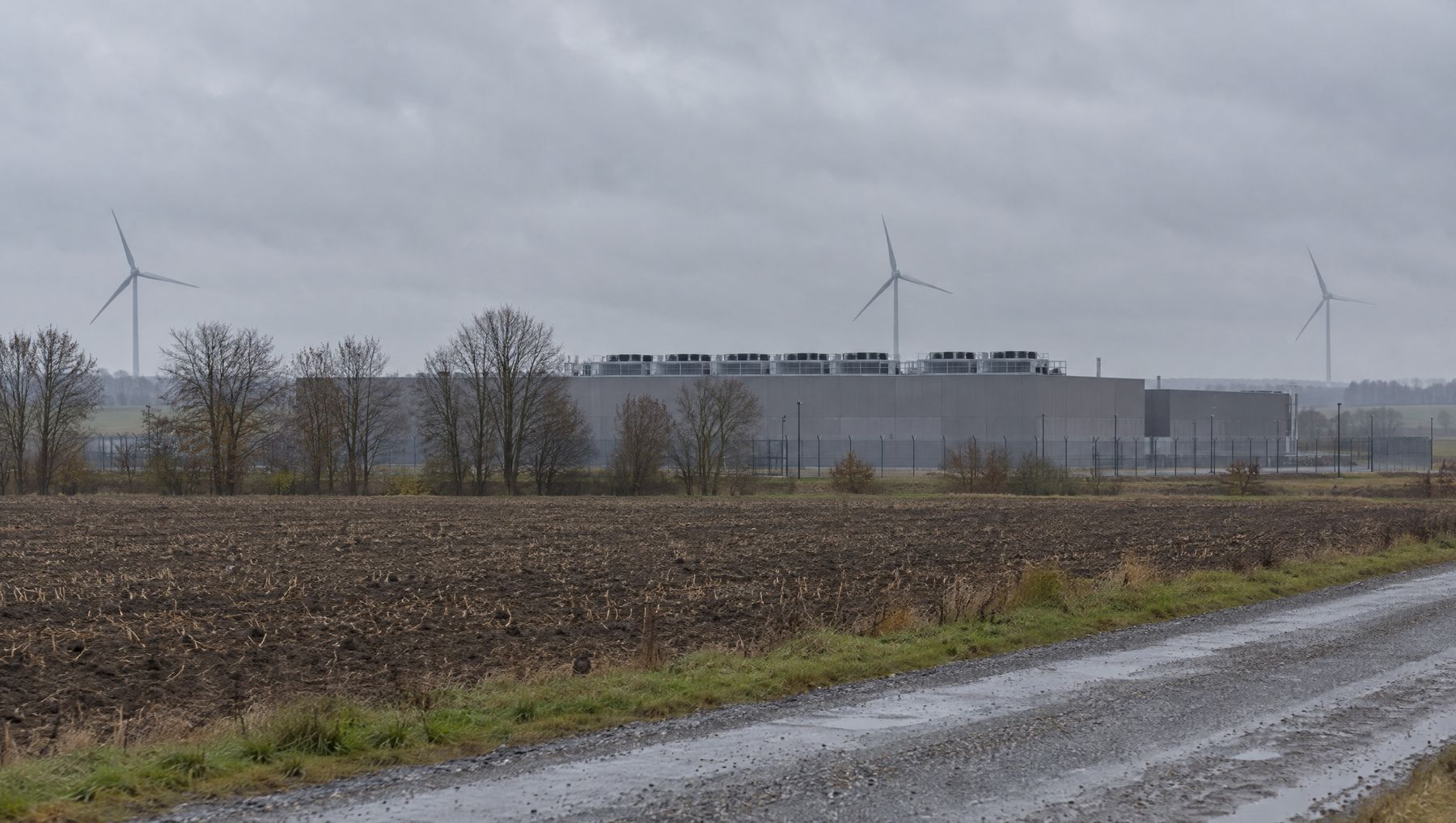 Lav grå datacenterbygning bag en pløjemark med vindmøller i baggrunden