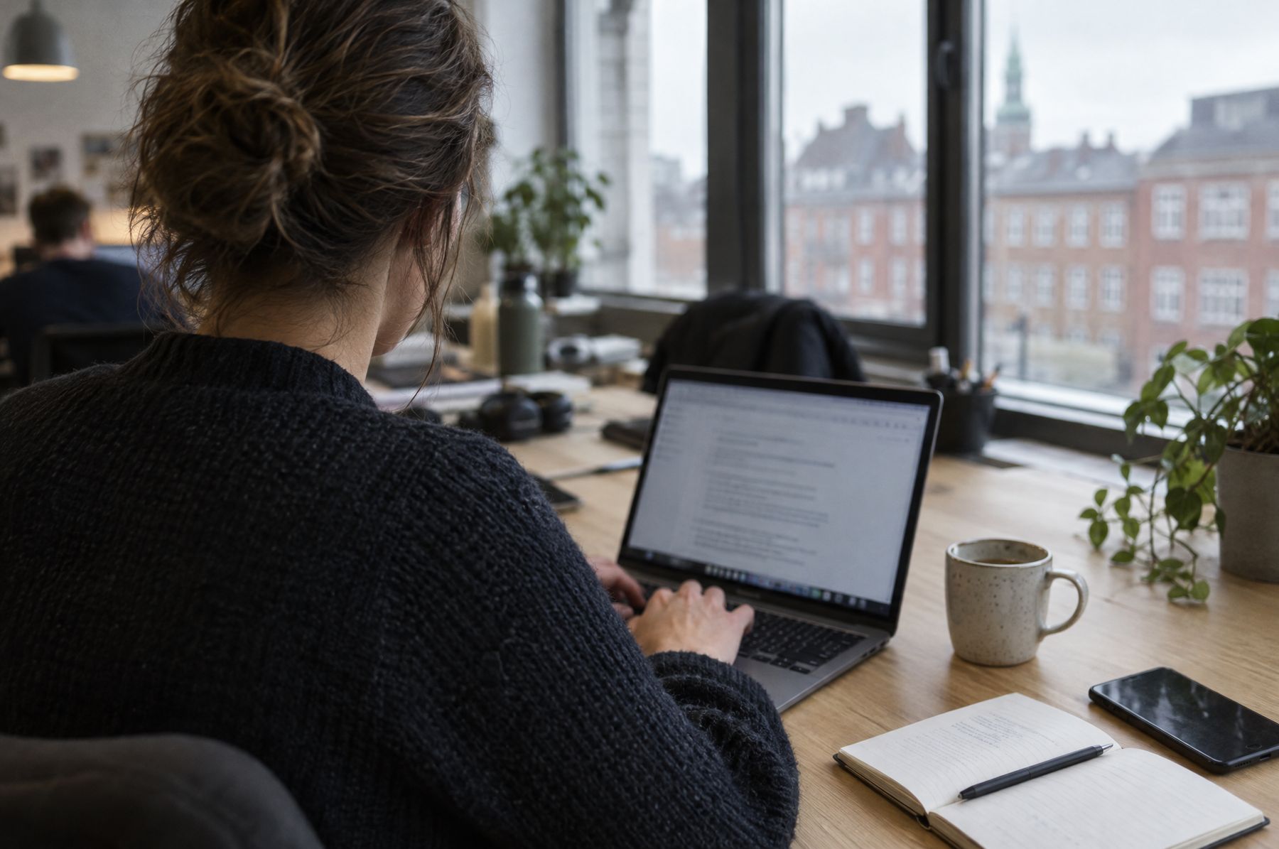 Person set bagfra, der arbejder ved en bærbar computer på et kontor med udsigt til røde murstensbygninger